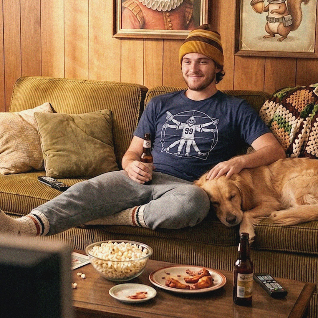 Man in a Solid Threads football graphic tee lounges on a retro corduroy couch with his dog, beer, popcorn and wings on game day.