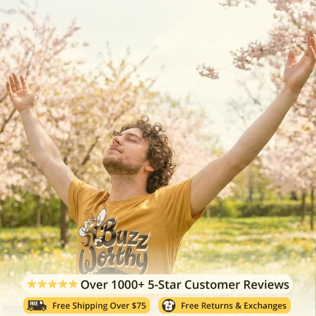 Man with arms outstretched in a cherry blossom garden, wearing a yellow 'Buzz Worthy' shirt.