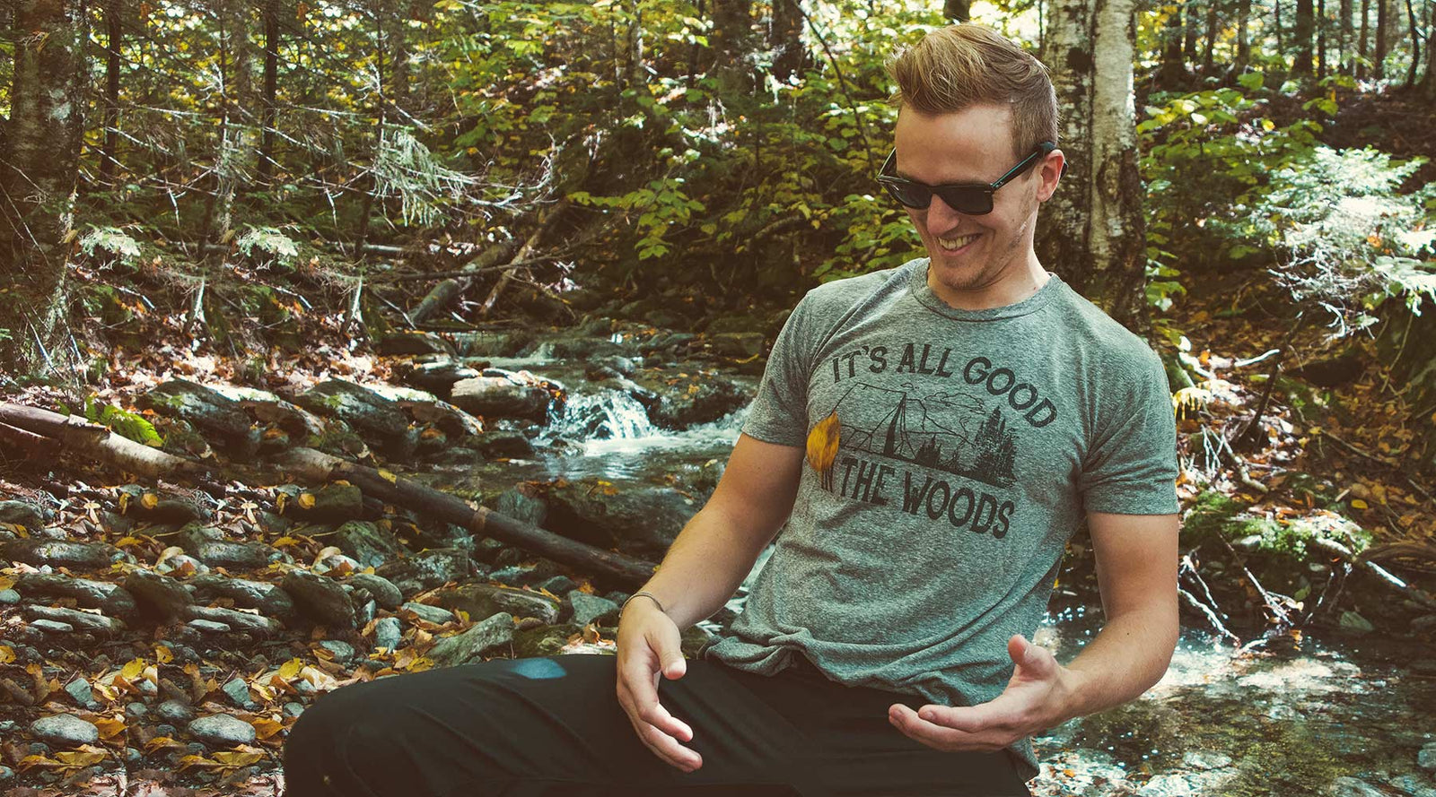 Man wearing a vintage-inspired Solid Threads graphic tee catches a falling autumn leaf beside a wooded stream.